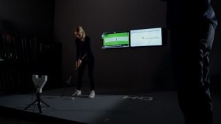 Image showing a woman putting in a dark putter fitting room in front of two TV screens on the wall.