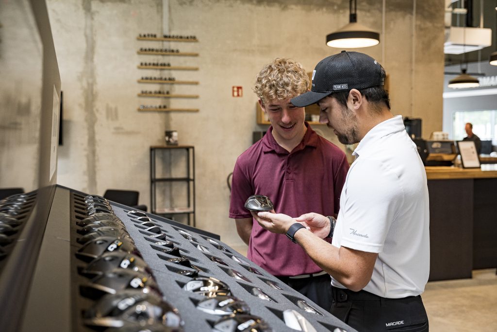 Image of a male golf club fitter showing a hybrid head to a young customer inside of an indoor fitting studio.