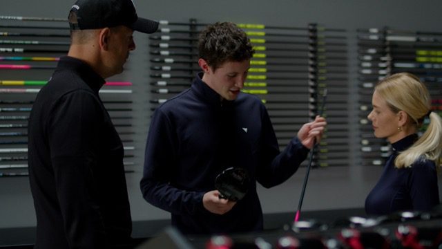 Image of a young club fitter explaining the components of a driver to a male and a female customer in front of a colorful shaft display.